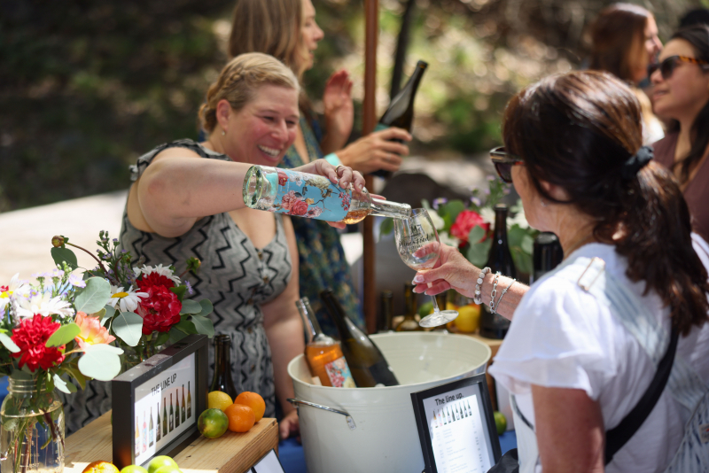 Smiling woman pours white wine into the photographer's glass in a wooded landscape