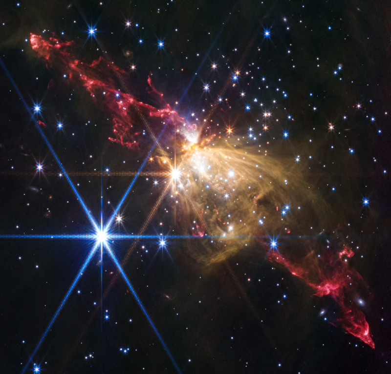A colorful image of wispy red and yellow streaks and bright stars displaying the six points characteristic of the JWST's hexagonal mirrors
