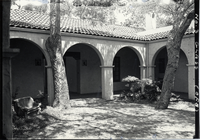 An old black-and-white photo of a corner of a courtyard, with heavy arches and tile roof. Two large oak trees are growing in the courtyard.