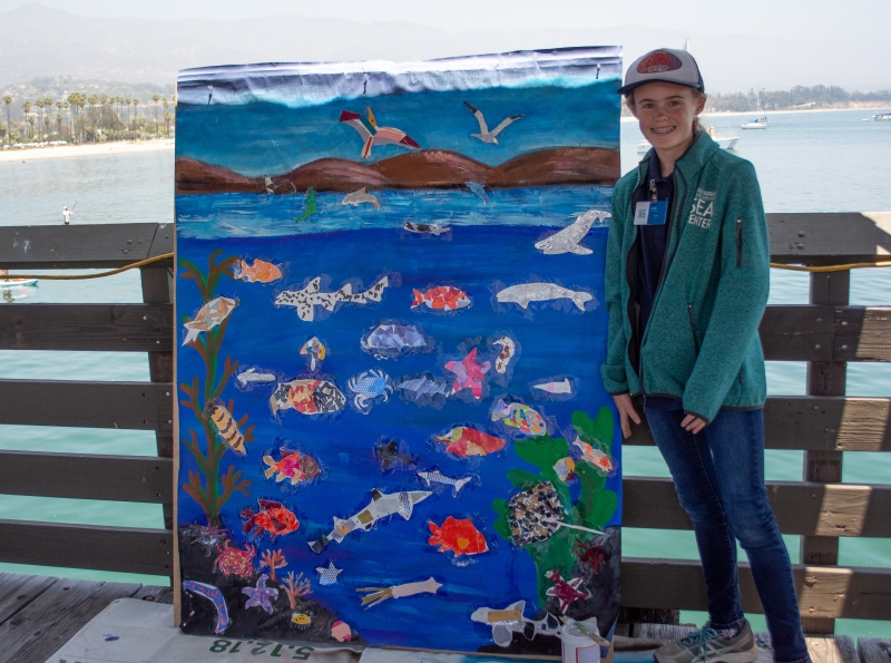 Lucy London as a young girl, standing at the Sea Center with the ocean in the background. She's next to a collage board as tall as her, covered with brightly colored paintings of marine life